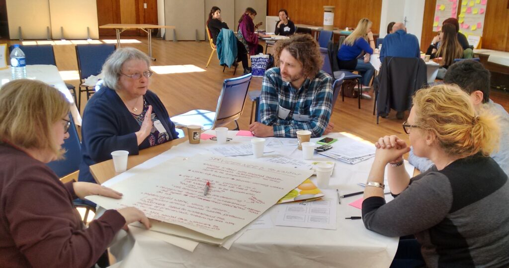 A group of five people at the People's Conference sitting around a table talking and writing. In the background are more people.