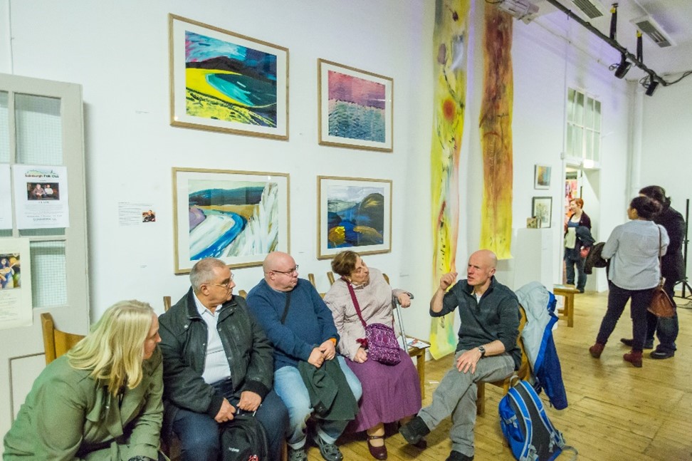 A group of five people sitting down talking within the exhibition, with art works in the background.