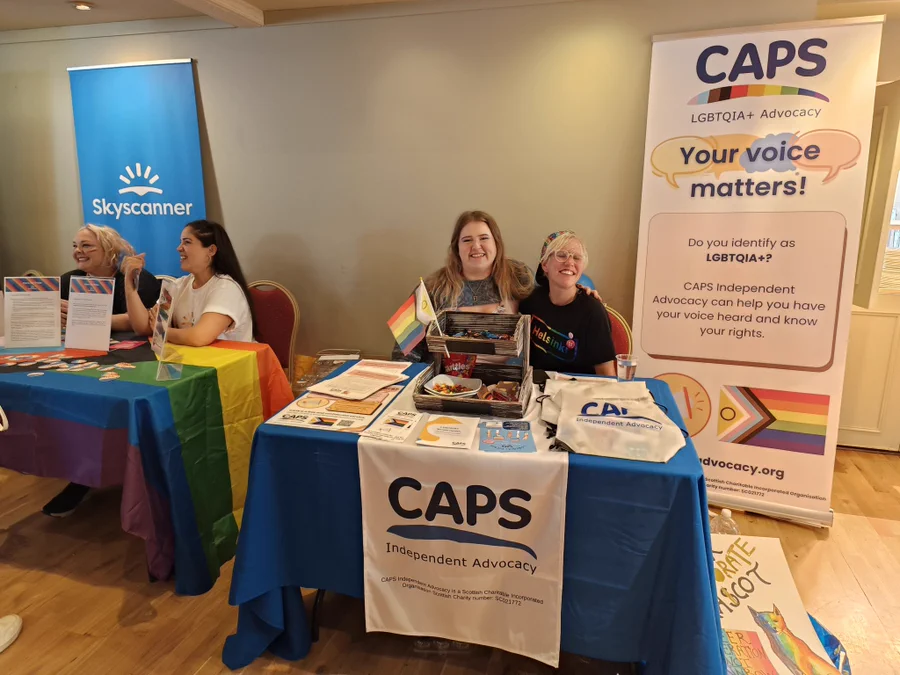 Malin and Ellis representing CAPS LGBTQIA+ work at a Pride event with CAPS banners and stall with leaflets.