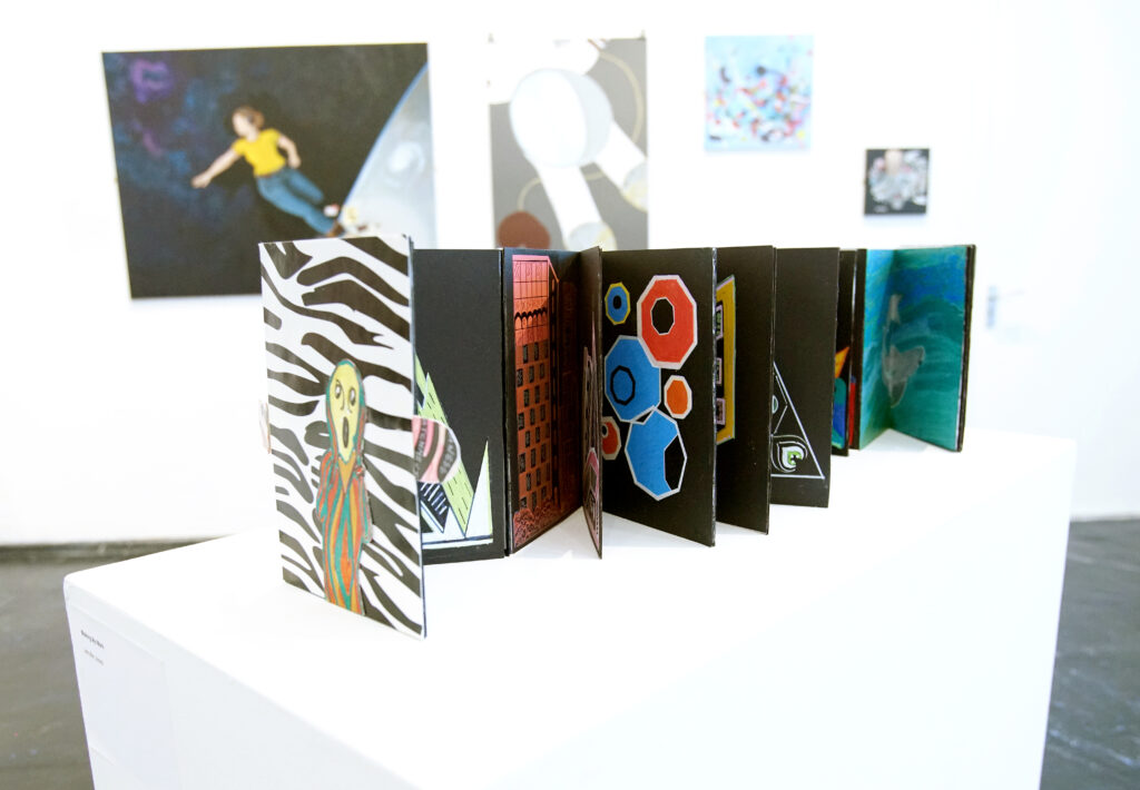 A photgraph of an artwork resting on a plinth. The artwork is a book opened out with brightly coloured patterns and paintings. In the bakcground are further artworks from the Out Of Sight Out Of Mind exhibition.