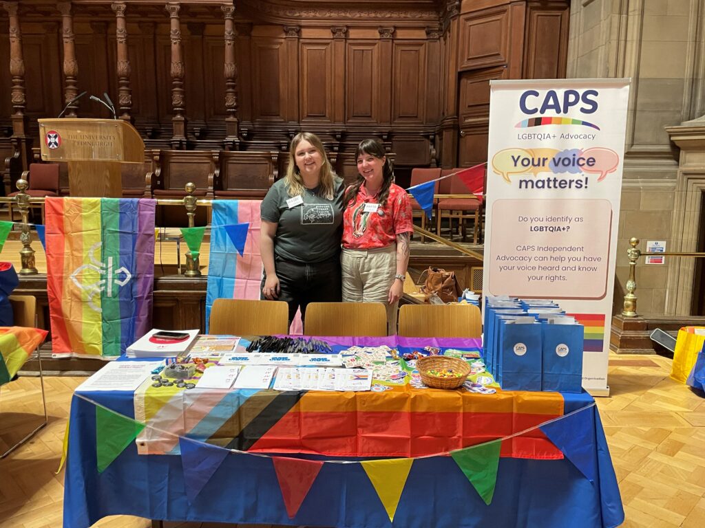 The CAPS stall at Pride in 2024, two people standing behind a table with pride flags and a banner that reads 'Your voice matters!'