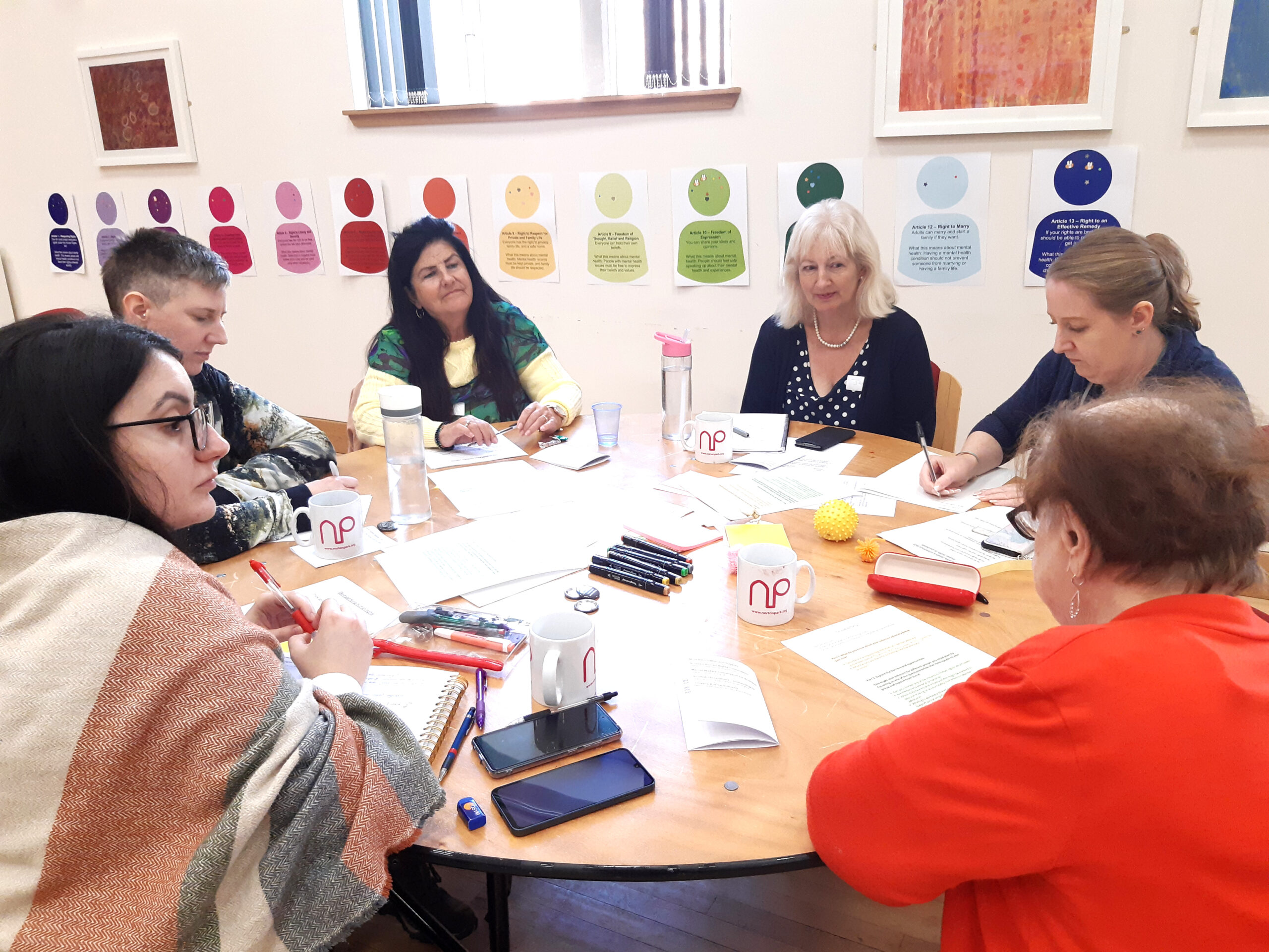 Six people sitting around a table discussing, on the table are pieces of paper and brightly coloured pens.